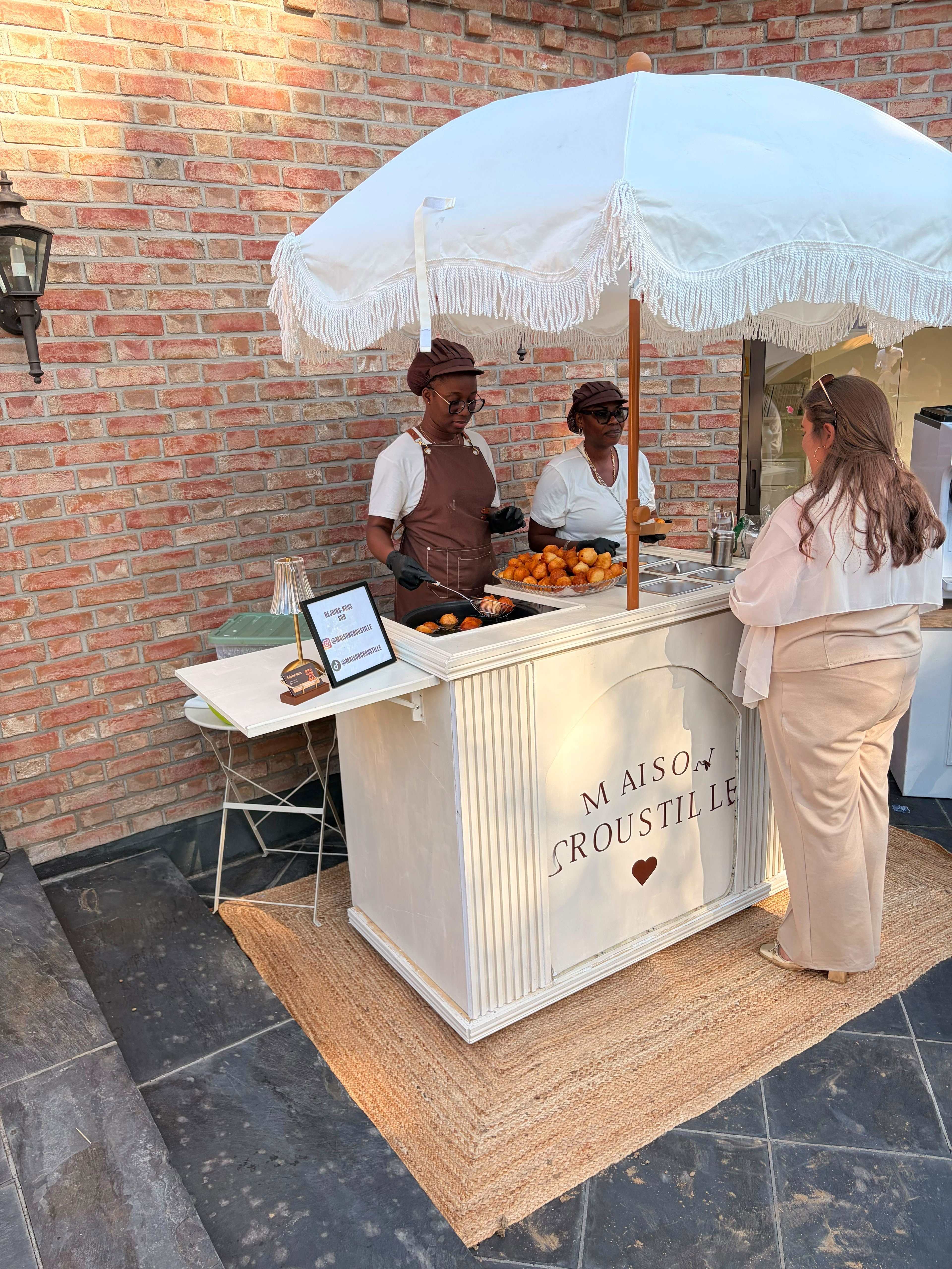 Deux hôtes Maison Croustille en tablier chocolat et casquette font dorer les mikatés au comptoir, sous le parasol à franges.