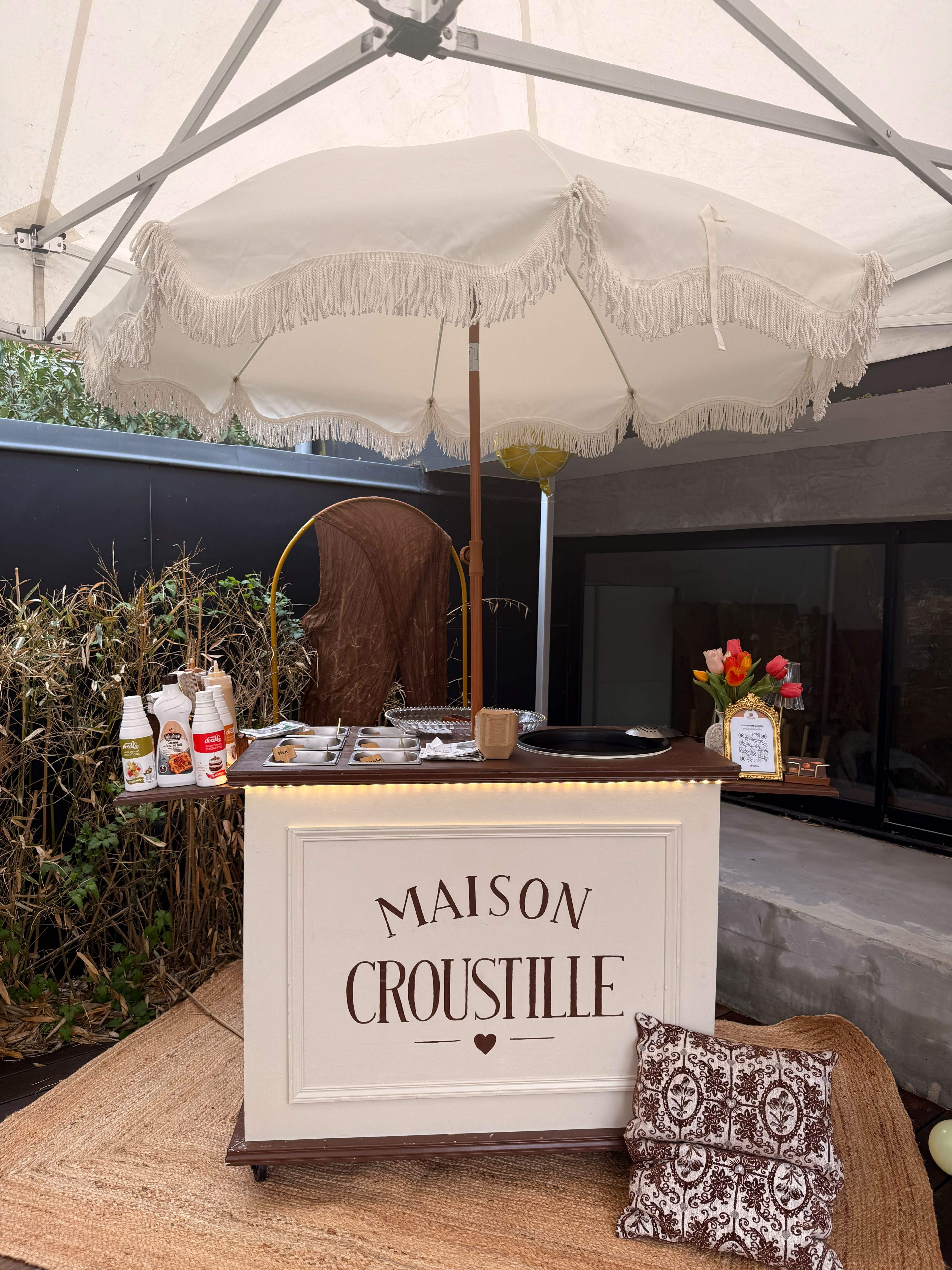 Le comptoir Maison Croustille installé sous une tente : parasol blanc à franges, panneau crème au nom de la maison, bouquet de tulipes et coussins.